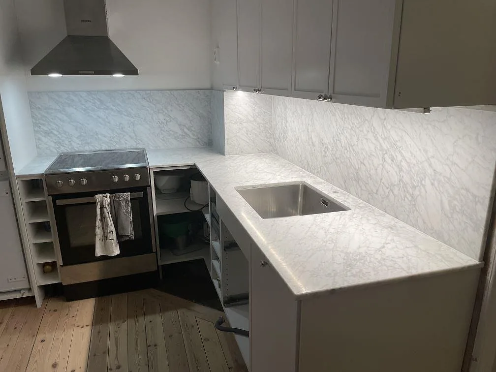 Matt Carrara C marmor som köksbänkskiva och stänkskydd i ett kök i Stockholm, med rostfri diskho och fristående induktionsspis mellan öppna köksstommar.