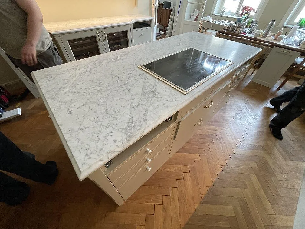 Fristående köksö i Carrara C marmor med kraftig diagonal grå ådring och infälld induktionshäll i ett kök i Stockholm.