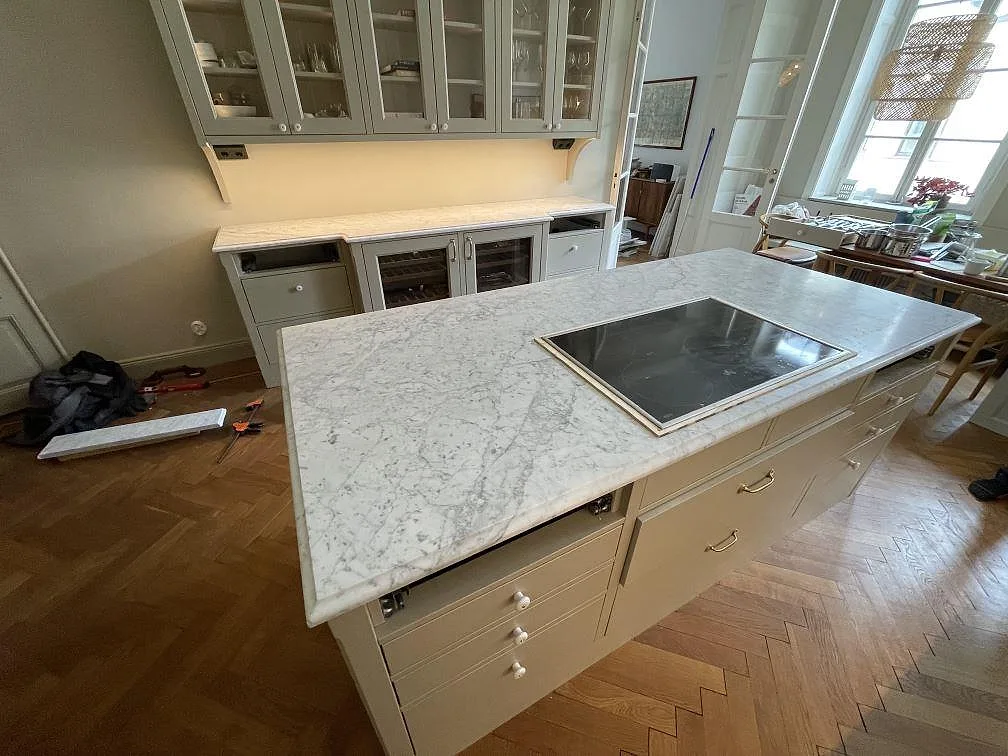 Köksö i vit Carrara C marmor med induktionshäll och glasvitrinskåp i bakgrunden i ett kök i Stockholm.