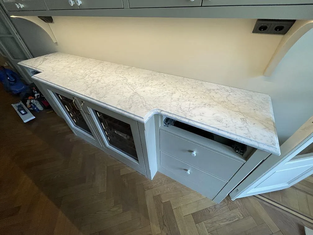 Bänkskiva i Carrara C marmor med fjäderlik grå ådring och glasvitrinskåp längs väggen i ett kök i Stockholm.
