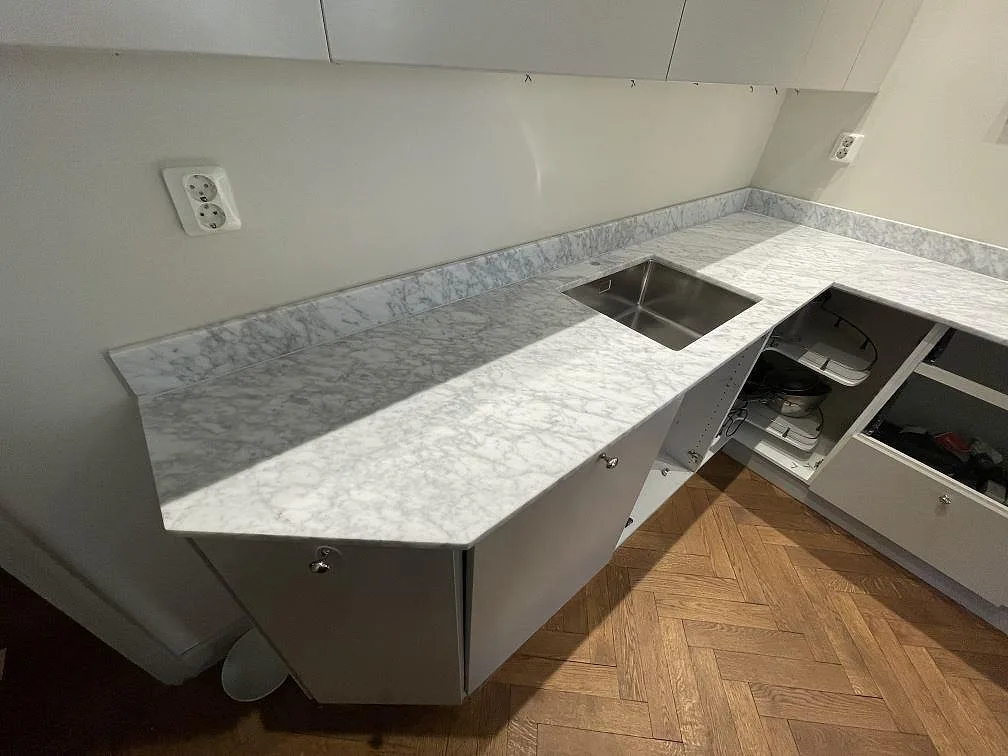 Matt Carrara C-marmor med mjuk grå ådring längs en lång köksbänk med underlimmad rostfri diskho och låg stänkkant i Stockholm.