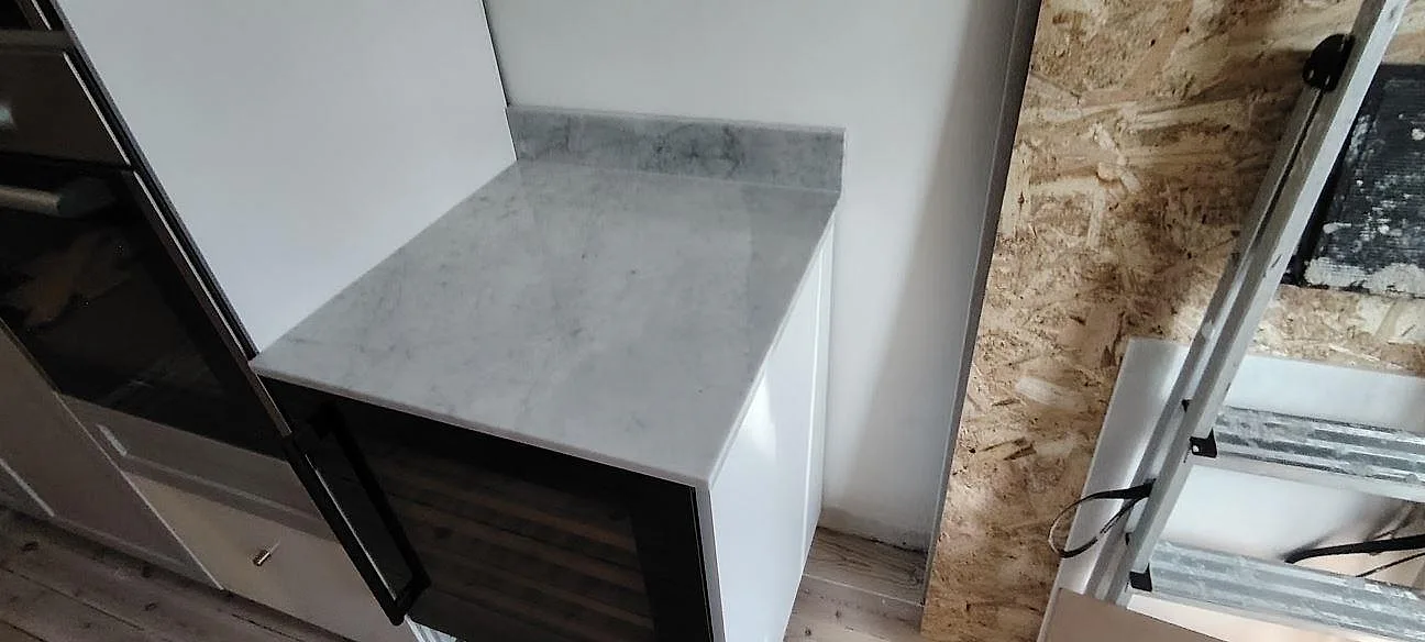 Sidoyta i polerad Carrara C marmor med låg stänkkant intill högskåp i ett kök under montering i Sundbyberg.