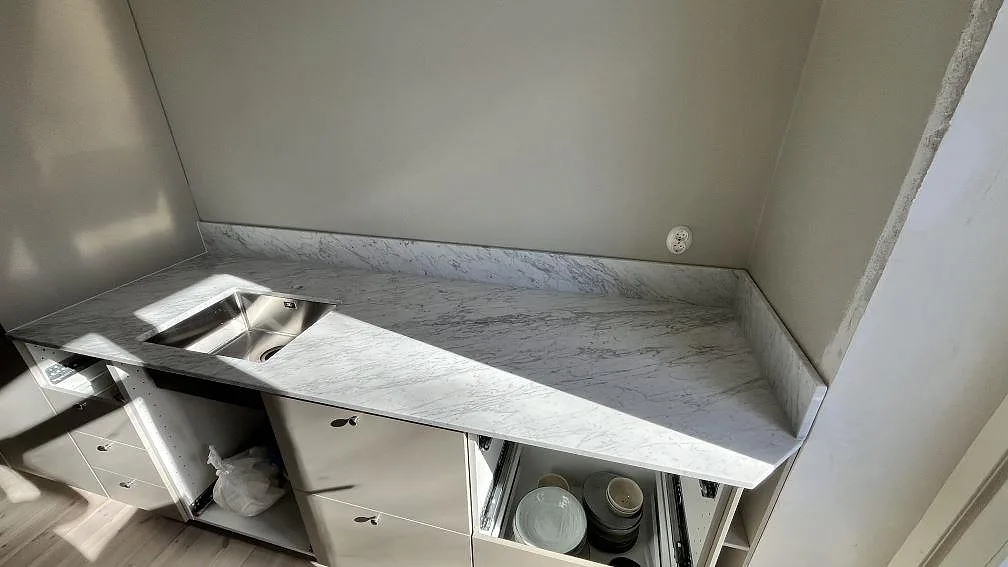 Matt köksbänkskiva i Carrara C marmor med rostfri diskho och låg stänkkant i ett kök i Sundbyberg.