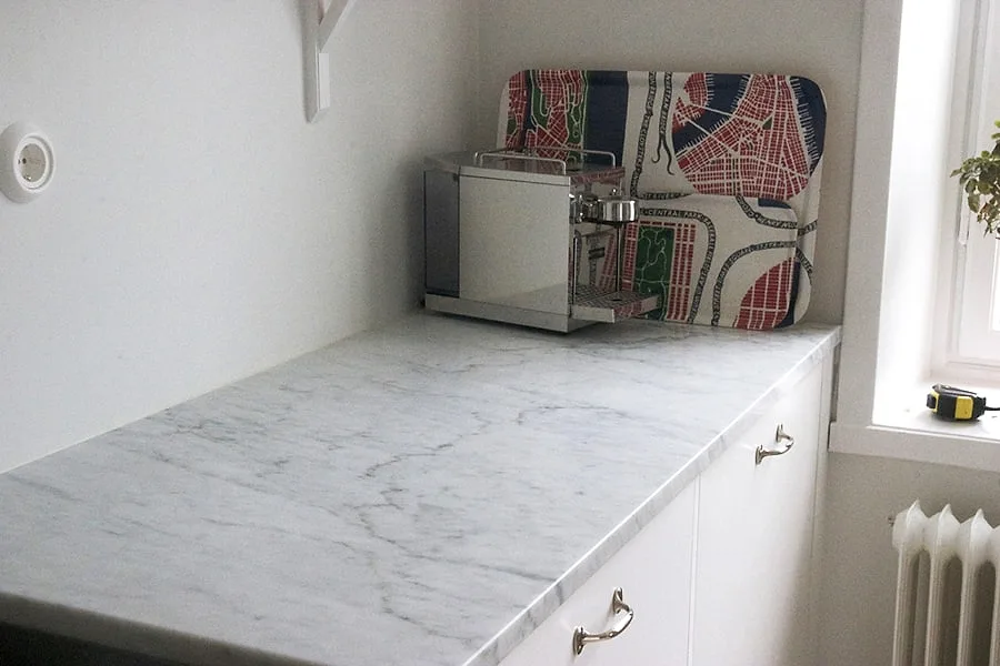 Köksbänkskiva i vit Carrara C-marmor, matt yta med grå ådring, monterad på vita köksluckor vid ett fönster i ett kök i Sverige.