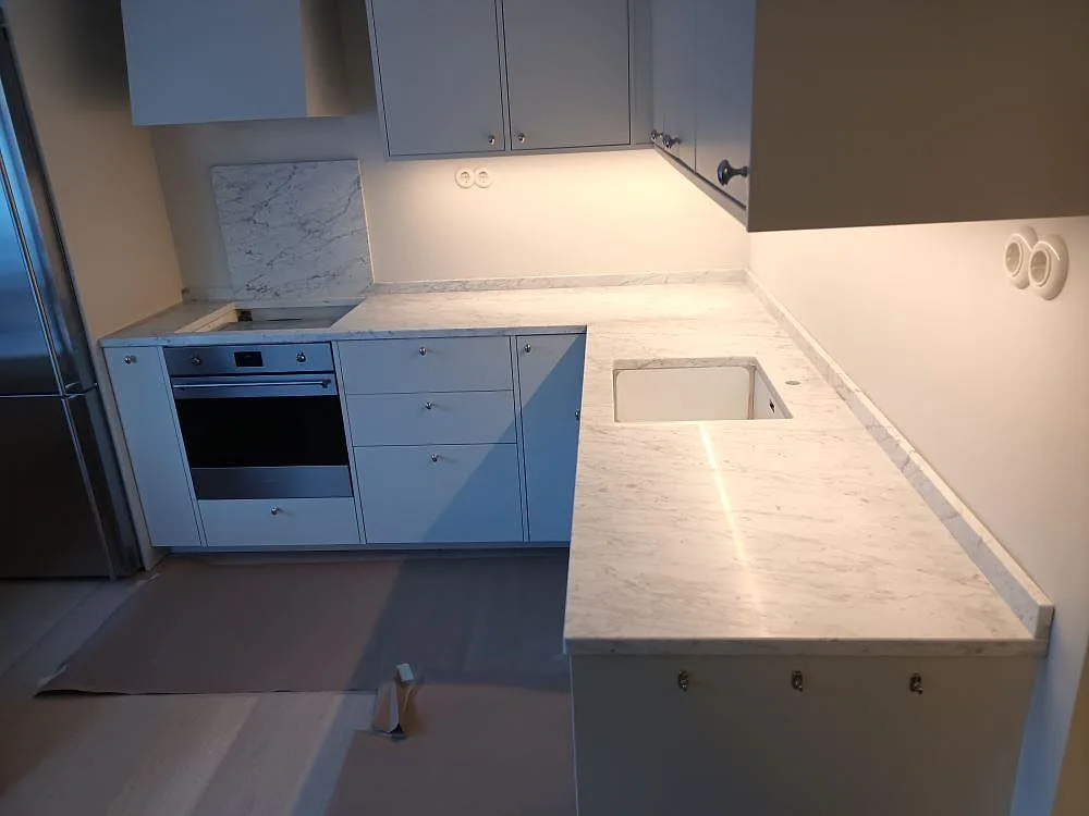 Köksbänkskiva i vit Carrara C-marmor med grå ådring, matt yta, med underlimmad diskho och uppvikt stänkskydd i ett L-format kök i Täby.