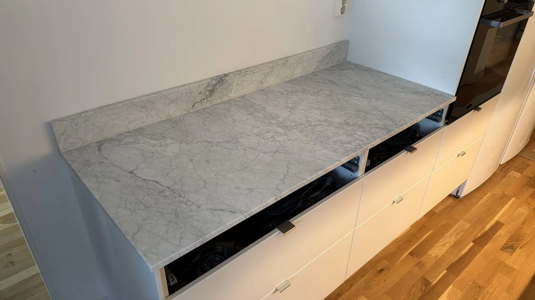 Rak köksbänkskiva i matt Carrara C marmor med uppvikt stänkkant och tydlig grå ådring vid högskåp i Täby.