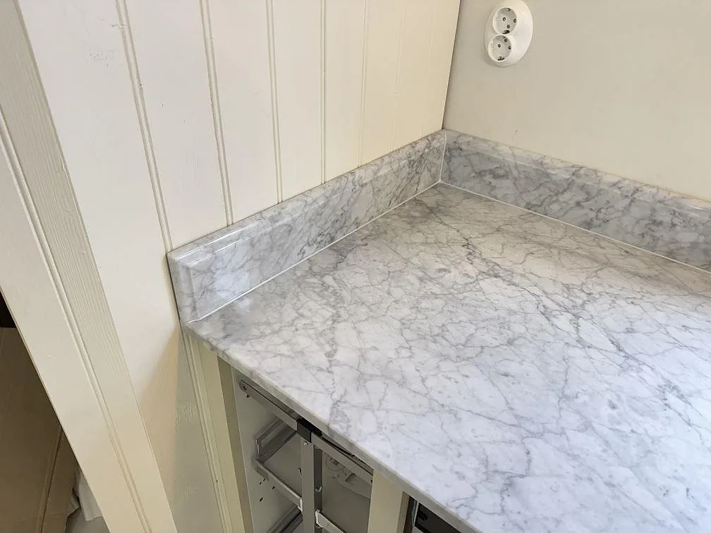 Hörn av polerad Carrara C marmorbänkskiva med låg stänkkant och öppna stommar under montering i Tenhult.
