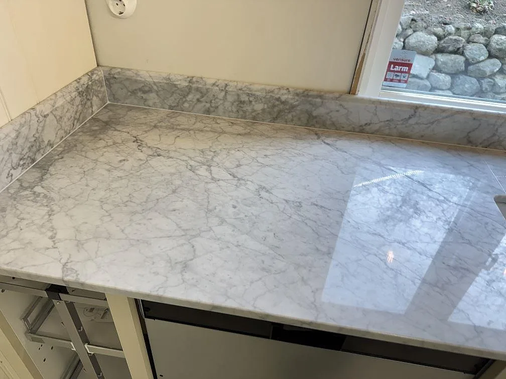 Polerad köksbänkskiva i Carrara C marmor med uppvikt stänkkant vid fönstret i ett kök i Tenhult.