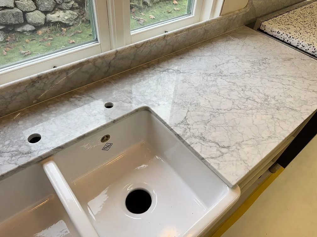 Carrara C marmor som polerad köksbänkskiva med vit diskho och stänkkant längs fönsterväggen i Tenhult.