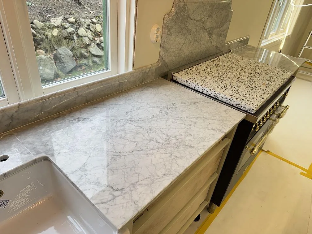 Köksbänkskiva i polerad Carrara C marmor med vit diskho, uppvikt stänkkant och spis under montering i Tenhult.