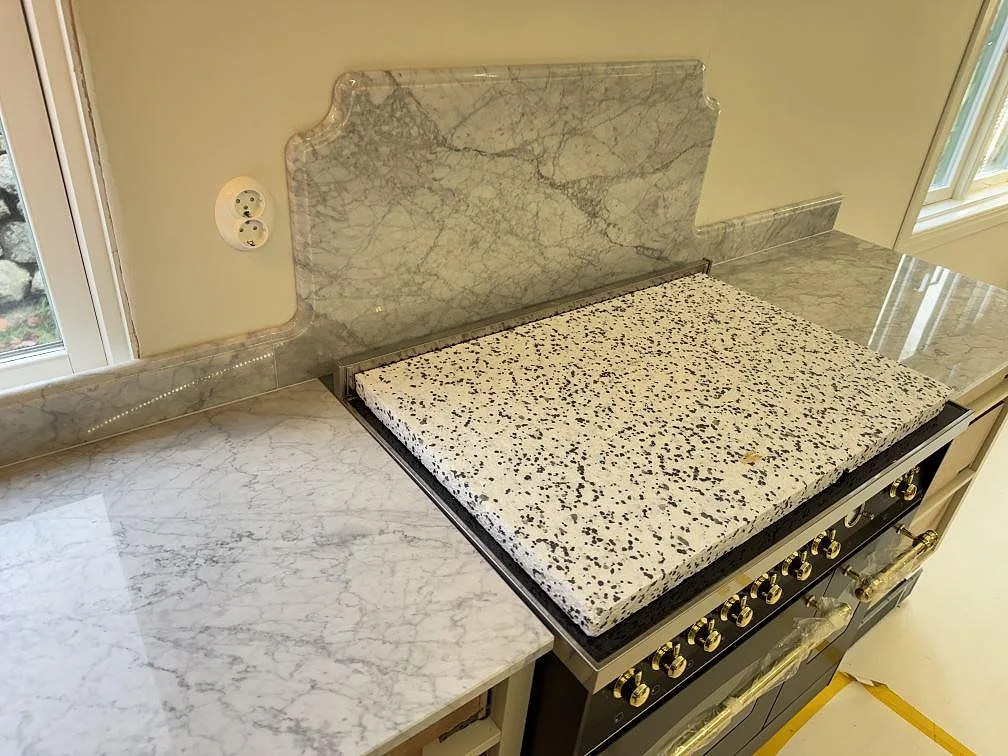 Formskuret stänkskydd i polerad Carrara C marmor bakom spisen och bänkskiva vid fönstret i Tenhult.