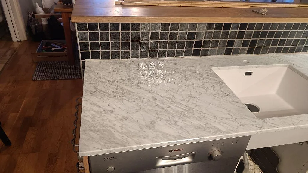Närbild på polerad Carrara C marmorbänkskiva med underlimmad diskho, diskmaskin och mörk mosaik i Timmersdala.