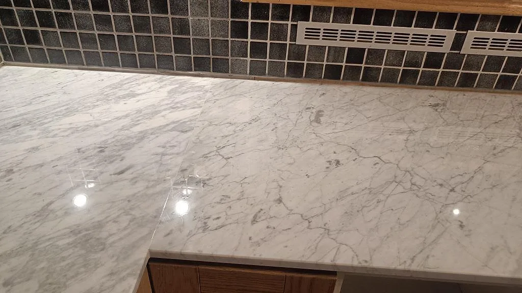 Polerad Carrara C marmorskiva över köksbänk med speglande yta, grå ådring och mörk mosaikvägg i Timmersdala.