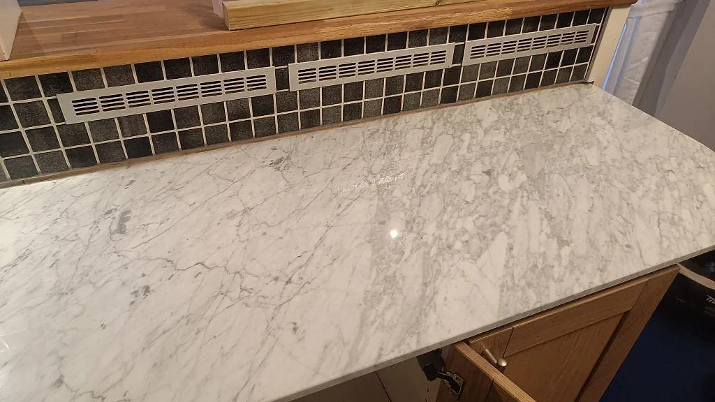 Lång arbetsyta i polerad Carrara C marmor med träinredning och mörkt mosaikstänkskydd i kök i Timmersdala.