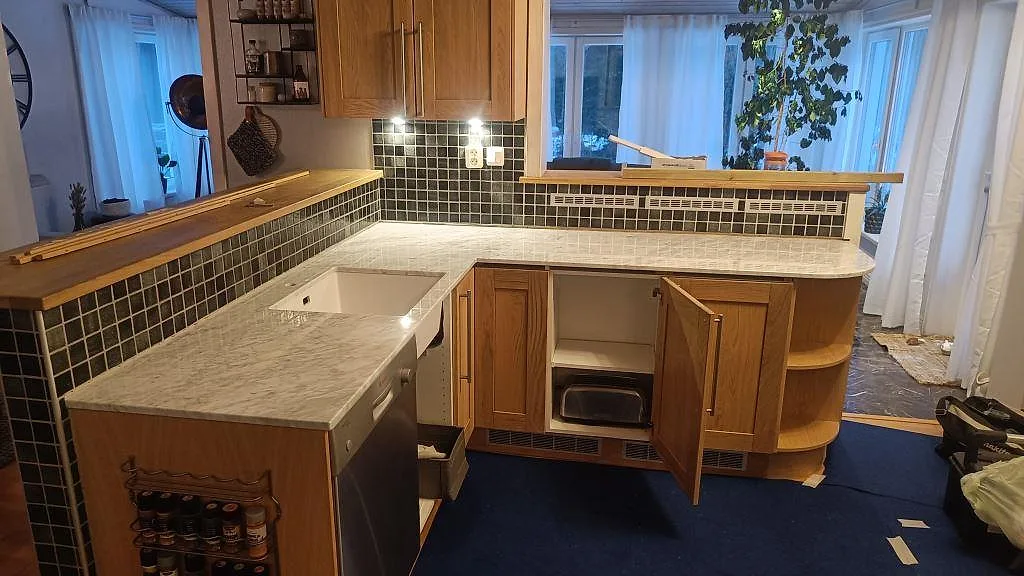 Polerad köksbänkskiva i Carrara C marmor med underlimmad vit diskho, mörkt mosaikkakel och träinredning i ett kök i Timmersdala.