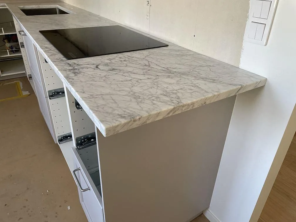 Köksbänkskiva i matt Carrara C marmor med induktionshäll och rostfri diskho i ett kök under montering i Torekov.