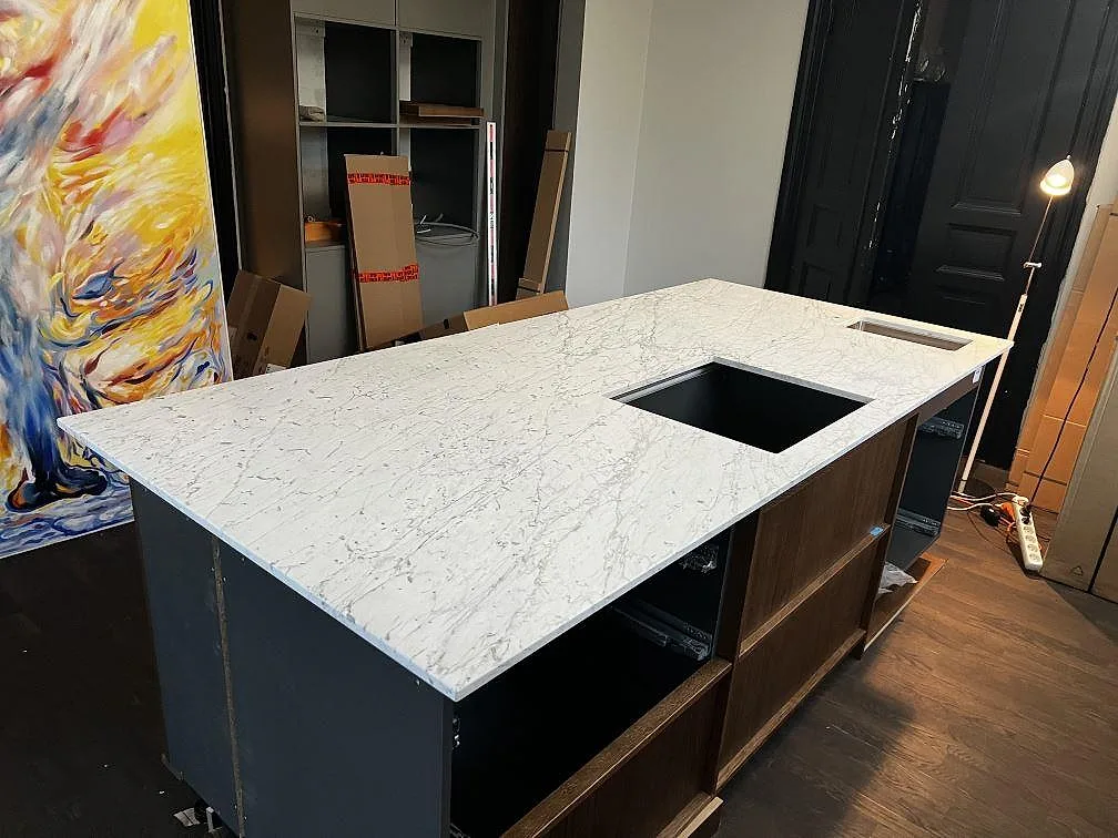 Stor köksö i Carrara C marmor med tydlig grå ådring, mörka stommar och färgstark abstrakt konst i ett kök i Trelleborg.