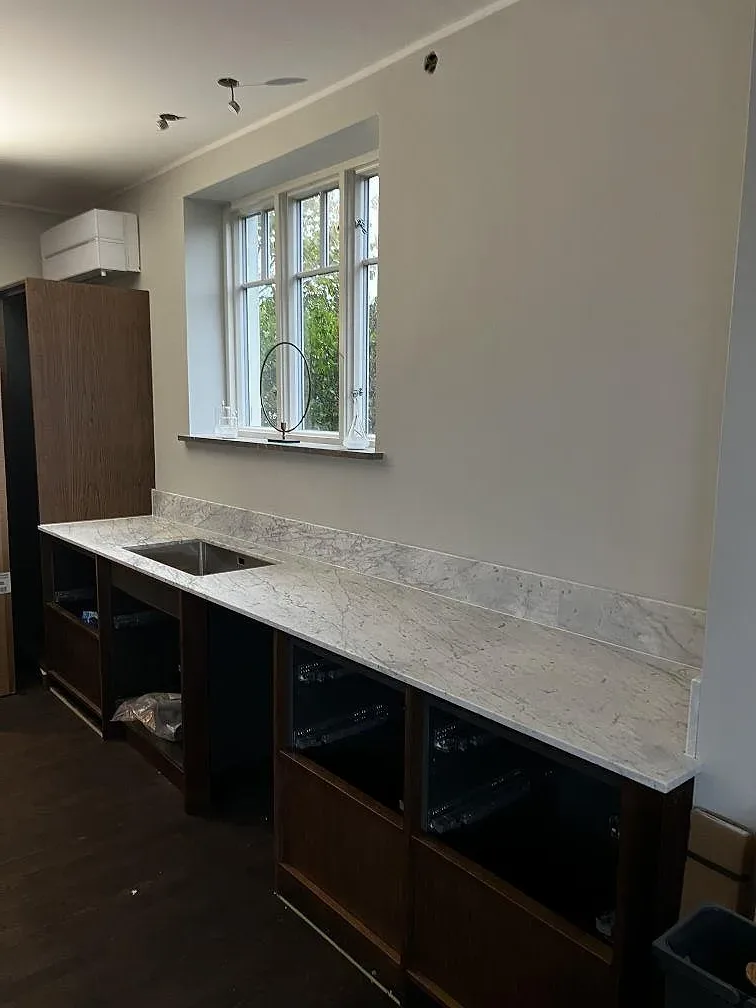 Lång köksbänkskiva i Carrara C marmor med uppvikt stänkkant, rostfri diskho och mörka stommar i Trelleborg.