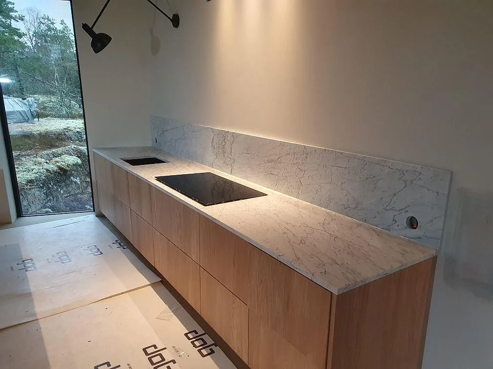 Köksbänkskiva i vit Carrara C marmor, matt, med underlimmad diskho och plats för induktionshäll samt uppvikt stänkskydd i ett kök i Trosa.