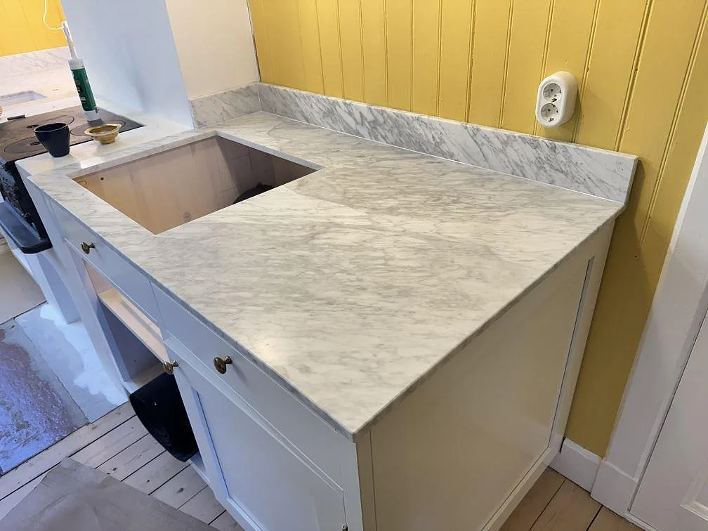 Köksbänkskiva i matt Carrara C marmor med uppvikt stänkkant och stor rektangulär öppning i ett kök i Uddevalla.