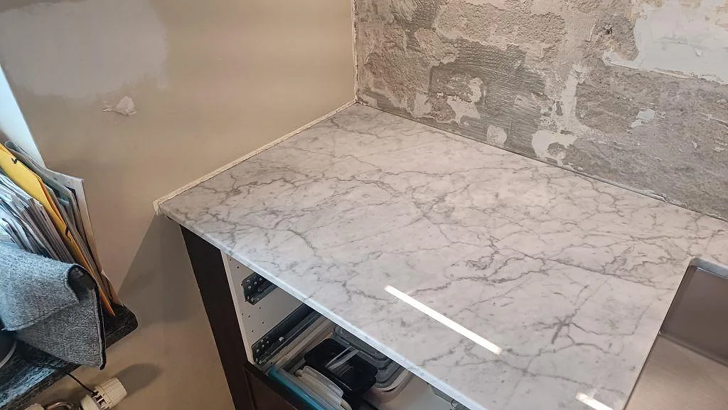 Hörnvy av polerad Carrara C marmorbänkskiva vid fönsteränden, med grå ådring och öppen köksstomme i Västra Frölunda.