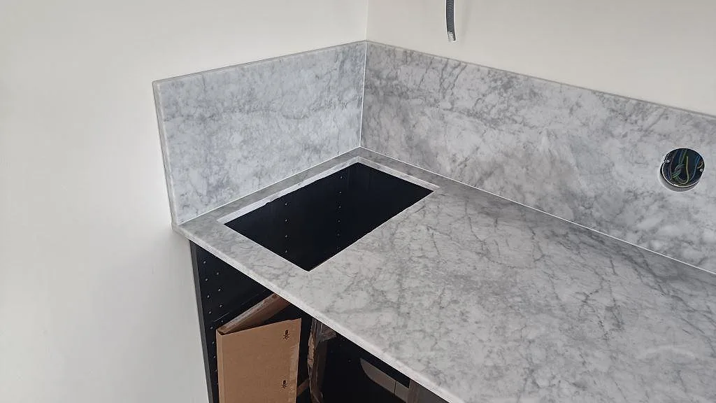 Hörnvy av köksbänkskiva i polerad Carrara C marmor med hällöppning och matchande stänkkant i Västra Karup.