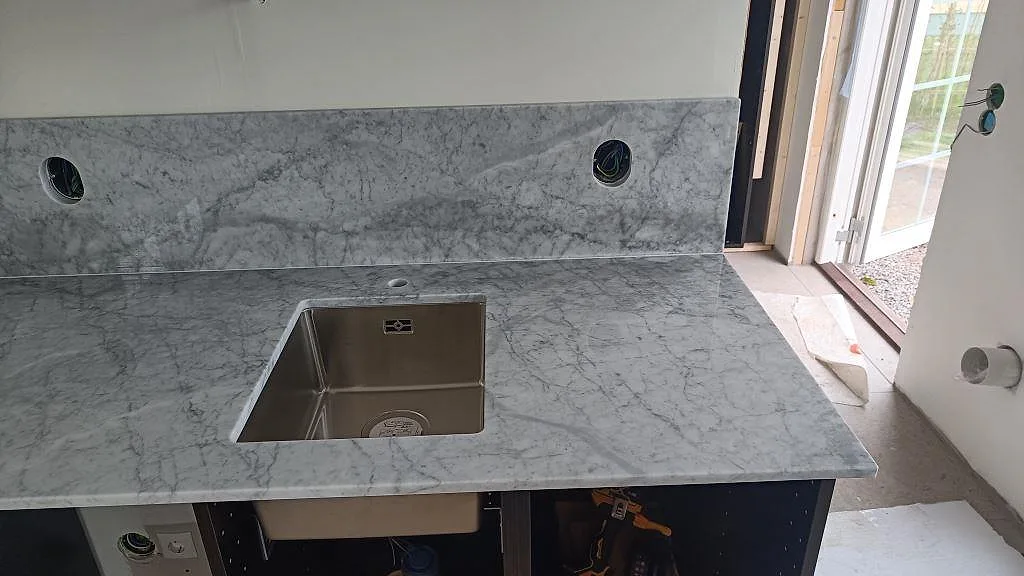 Diskhozonen i en köksbänkskiva av polerad Carrara C marmor med hög stänkkant och öppet monteringsskede i Västra Karup.
