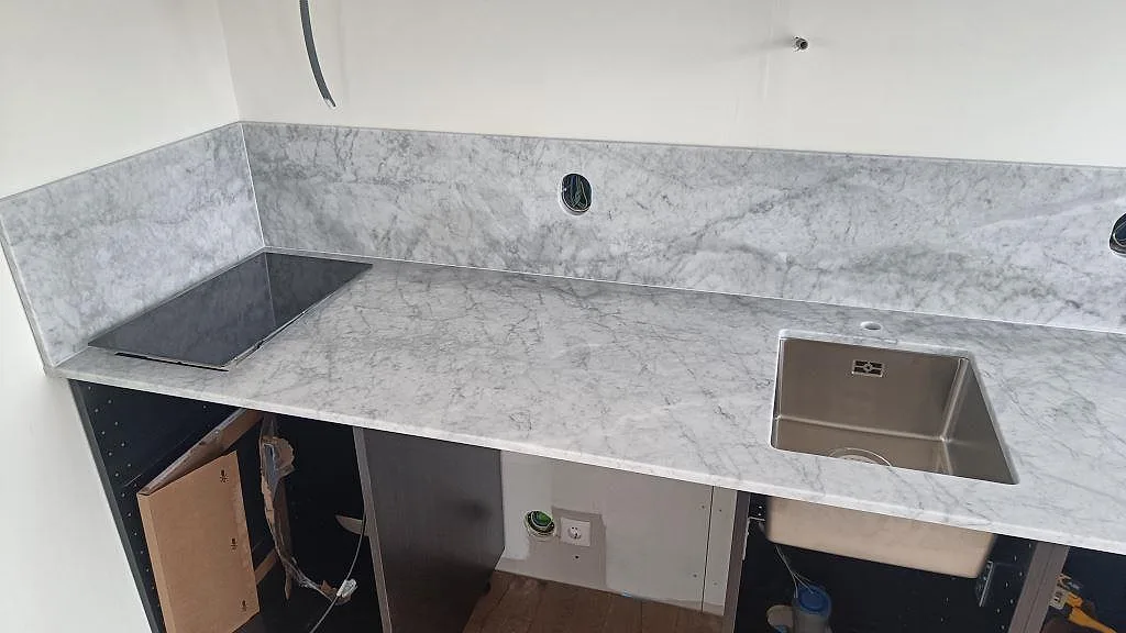 L-formad köksbänkskiva i polerad Carrara C med blank hällyta, diskho och uppvikt stänkkant i Västra Karup.