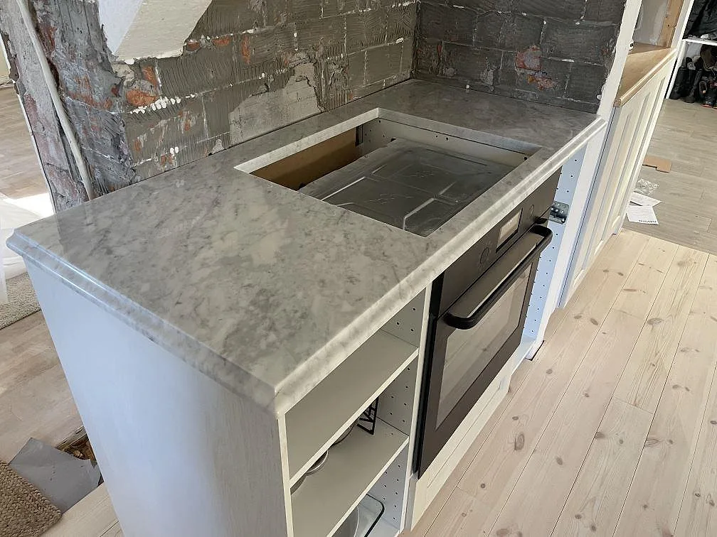 Köksbänkskiva i polerad Carrara C marmor med hällurtag ovanför ugn i ett kök under montering i Valbo.