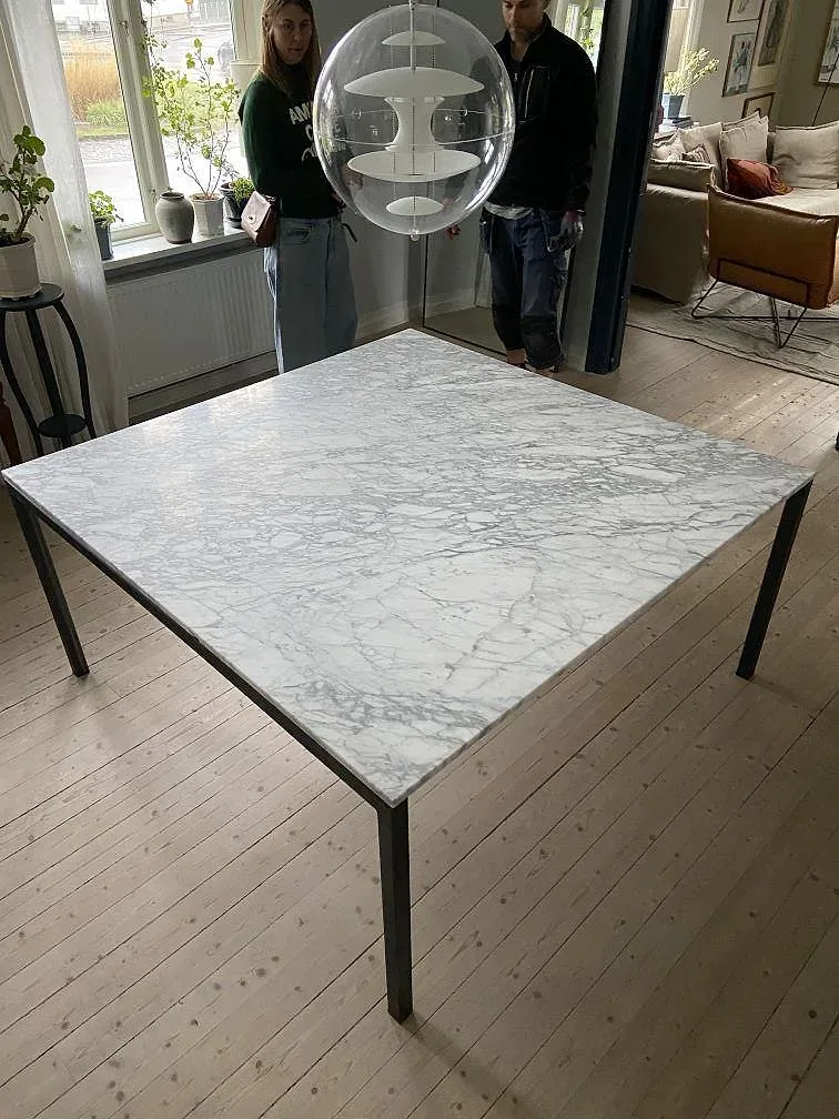Matbordsskiva i vit Carrara C marmor med grå ådring, matt yta, monterad på svart underrede i ett ljust vardagsrum i Varberg.