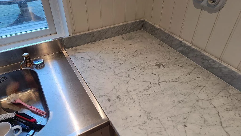 Carrara C marmorbänk i hörn med uppvikt marmorstänkkant och rostfri diskho i ett kök i Vejbystrand.
