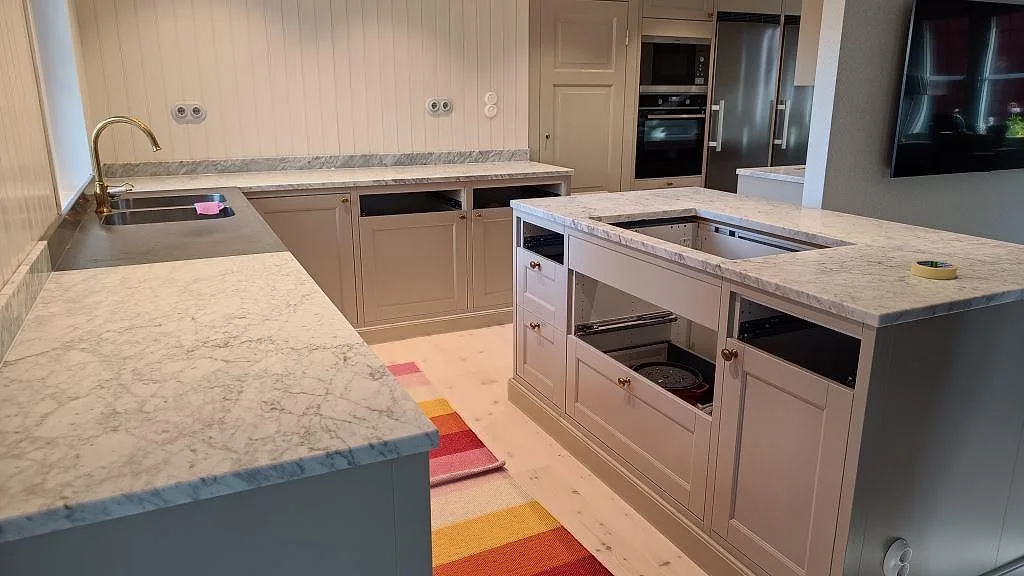 Köksbänkskivor i Carrara C vit och grå naturmarmor med matt yta, köksö och perimeterbänk med marmorstänkkant och rostfri diskho i ett kök i Vejbystrand.