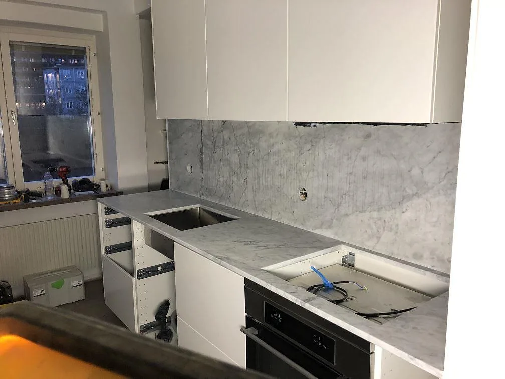 Köksbänkskiva och stänkskydd i vit Carrara CD marmor med grå ådring i ett L-format kök i Helsingborg, fotograferat under montering med underlimmad diskho och fönster.
