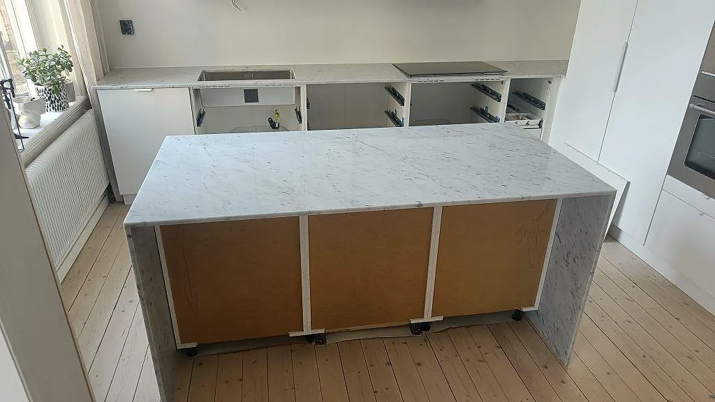 Köksbänkskiva och köksö i vit Carrara C marmor, polerad, med induktionshäll och underlimmad diskho i ett kök under montering i Helsingborg.