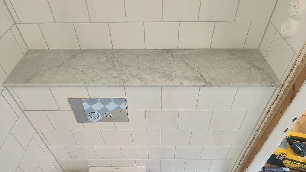 Smal marmorhylla i vit Carrara över wc:t i ett kaklat badrum i Höllviken.