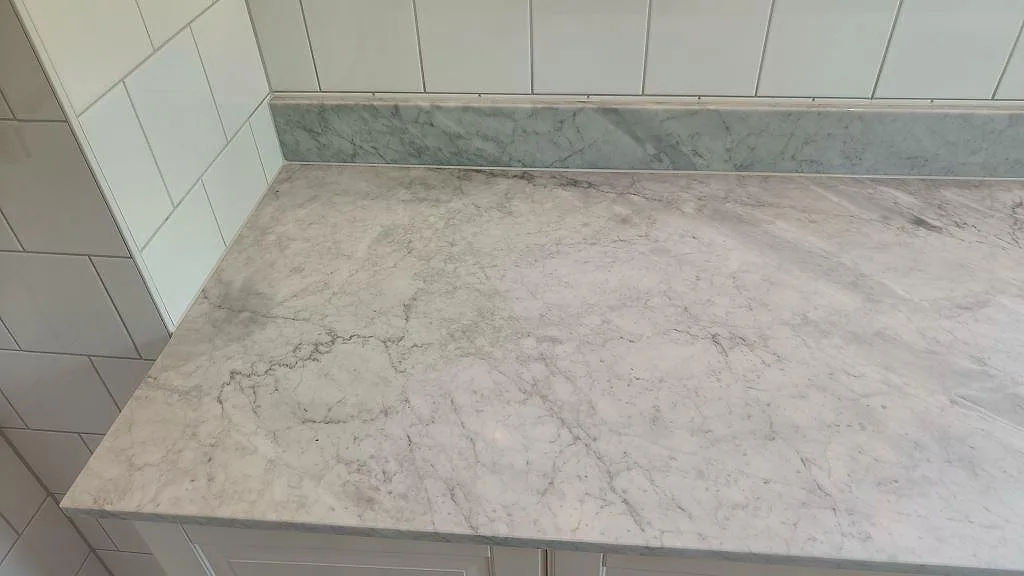 Matt Carrara-marmor med tydlig grå ådring som badrumsbänkskiva med uppvikt stänkkant i grågrön marmor i Höllviken.