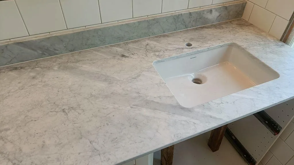Bänkskiva i vit Carrara-marmor med grå ådring i ett badrum i Höllviken, med underlimmat porslinstvättställ från Duravit och uppvikt stänkkant i grågrön marmor.