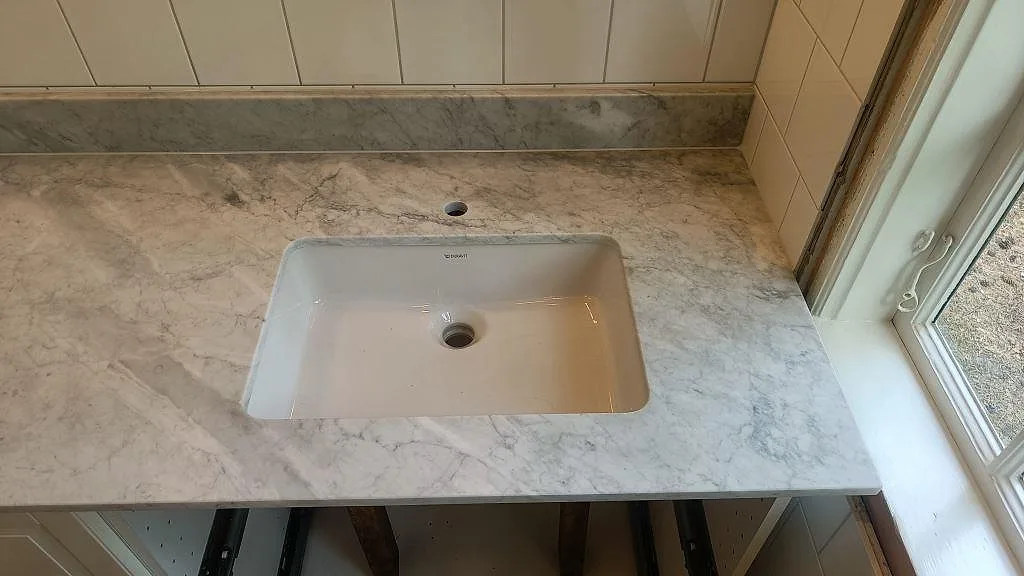Närbild på underlimmat tvättställ i ett badrum med bänkskiva i matt Carrara-marmor och kontrasterande grågrön marmorstänkkant i Höllviken.