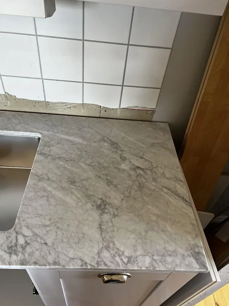 Detalj av matt Carrara C marmor vid diskhon, med vit kakelvägg och mässingshandtag i köket i Hunnebostrand.