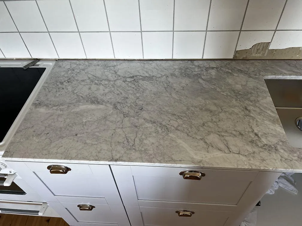 Matt köksbänkskiva i Carrara C marmor med synlig ådring, vitt kakel, rostfri diskho och hällsida i Hunnebostrand.