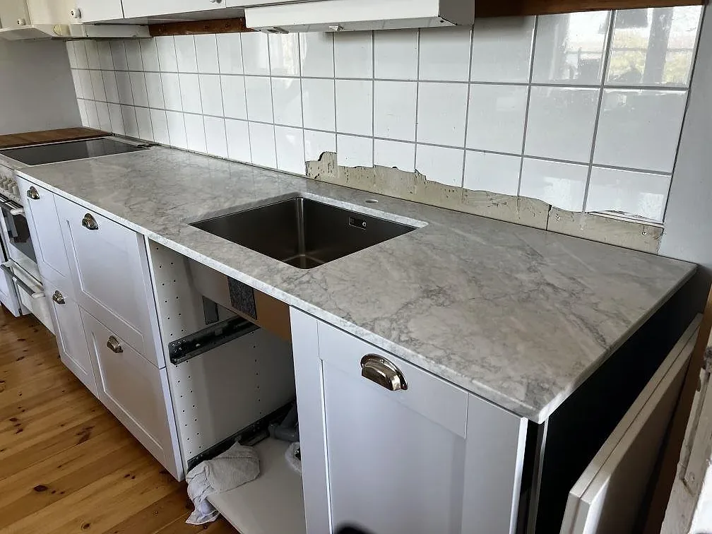 Köksbänkskiva i matt Carrara C marmor med underlimmad rostfri diskho, vita luckor och mässingshandtag i Hunnebostrand.