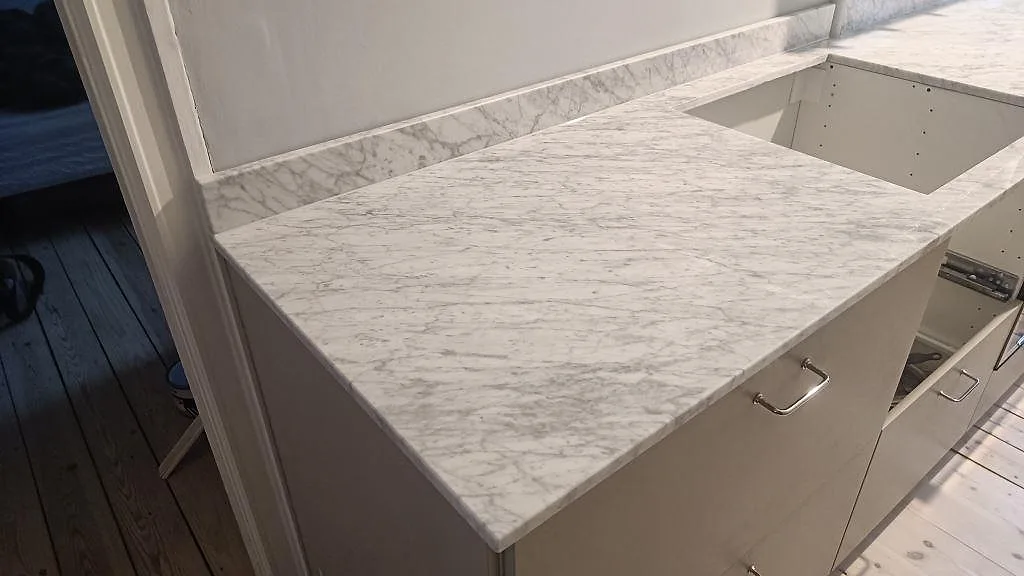 Närbild på polerad Carrara C köksbänkskiva med grå ådring, låg stänkkant och öppna vita köksstommar i København S.