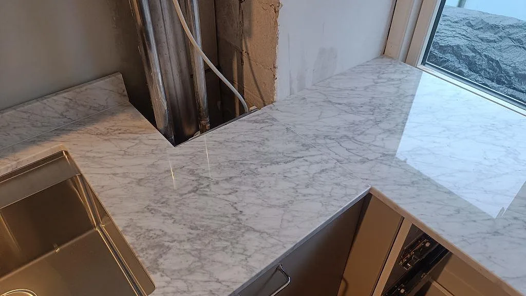 Hörnparti av polerad Carrara C köksbänkskiva vid fönster, med låg marmorstänkkant och pågående montering i København S.