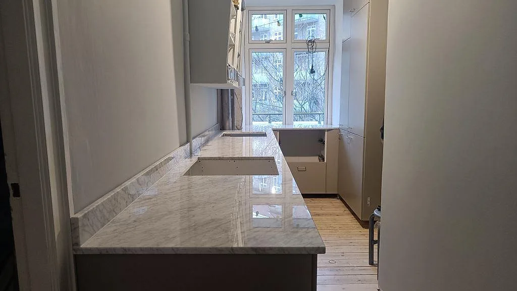 L-formad köksbänkskiva i polerad Carrara C marmor med låg stänkkant och köksö i ett kök under montering i København S.