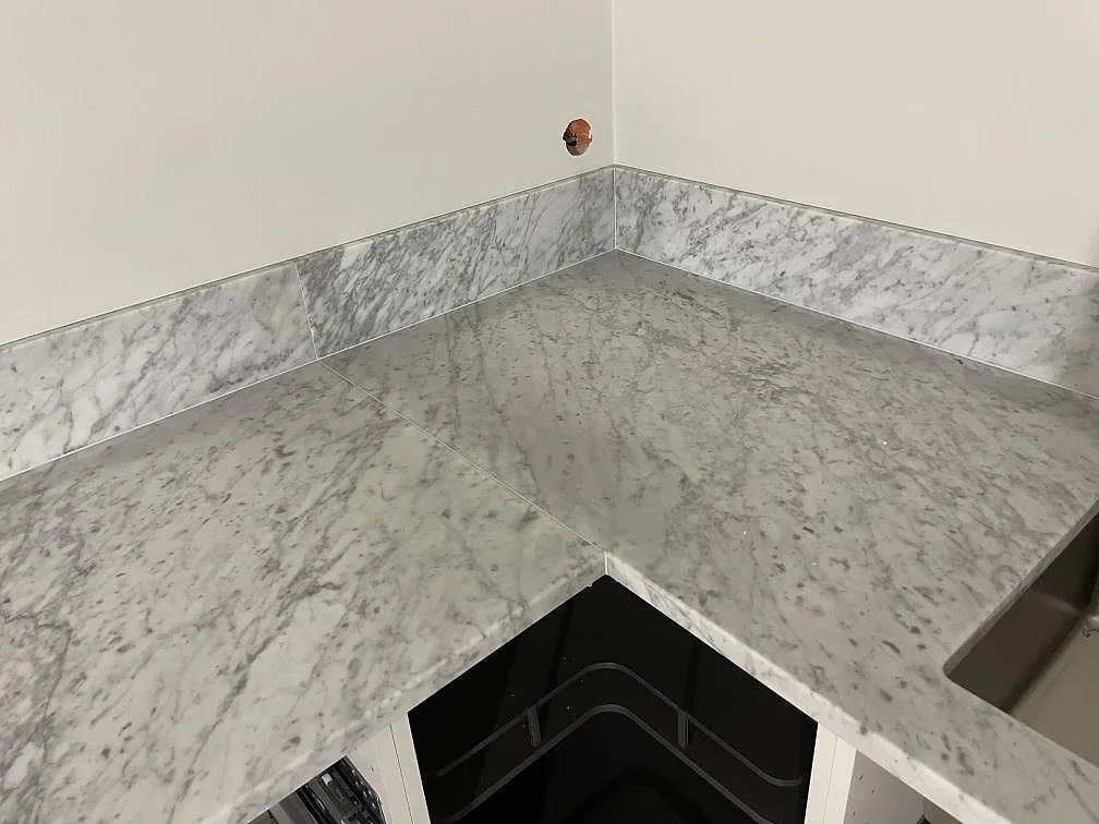 Hörnparti i matt Carrara C-marmor med grå ådring, stänkskydd och anslutning mot diskhon i Lund.
