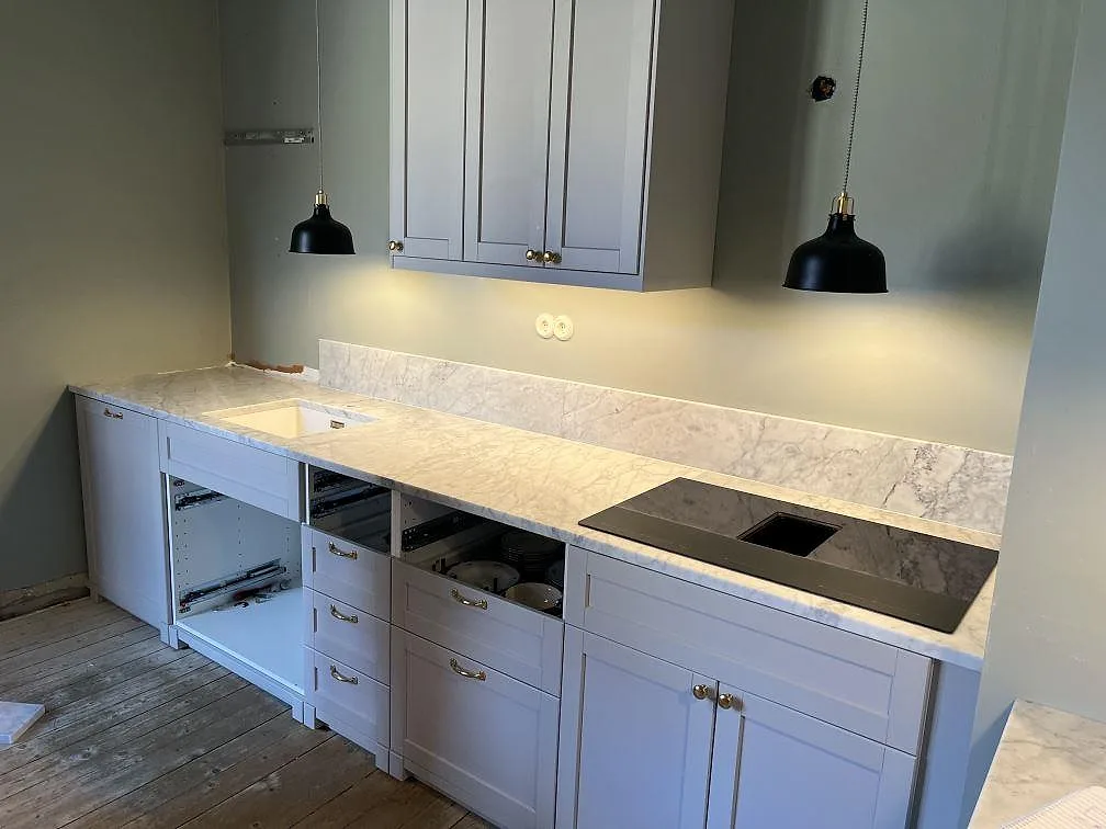 Köksbänkskiva i vit Carrara CD marmor, matt, längs en köksrad i grå luckor med underlimmad diskho, induktionshäll och uppvikt stänkskydd i Malmö.
