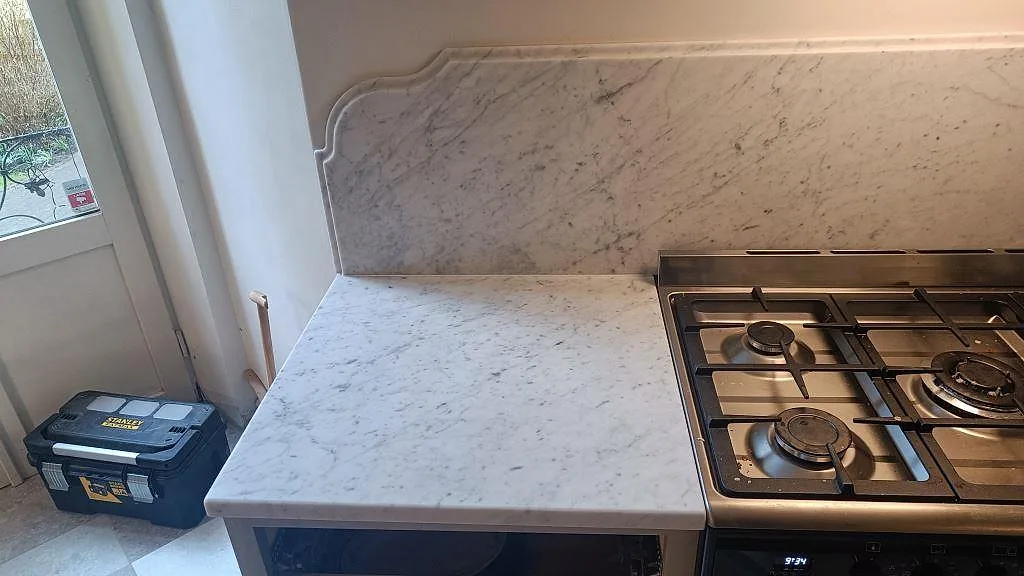 Vänster bänkdel i matt Carrara C marmor vid fönster och fristående gasspis, med profilerad stänkkant i ett Malmö kök.