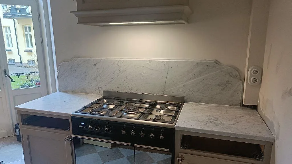 Köksbänkskiva i matt Carrara C marmor med uppvikt stänkkant, fristående gasspis, vita skåp och fönster i ett kök i Malmö.