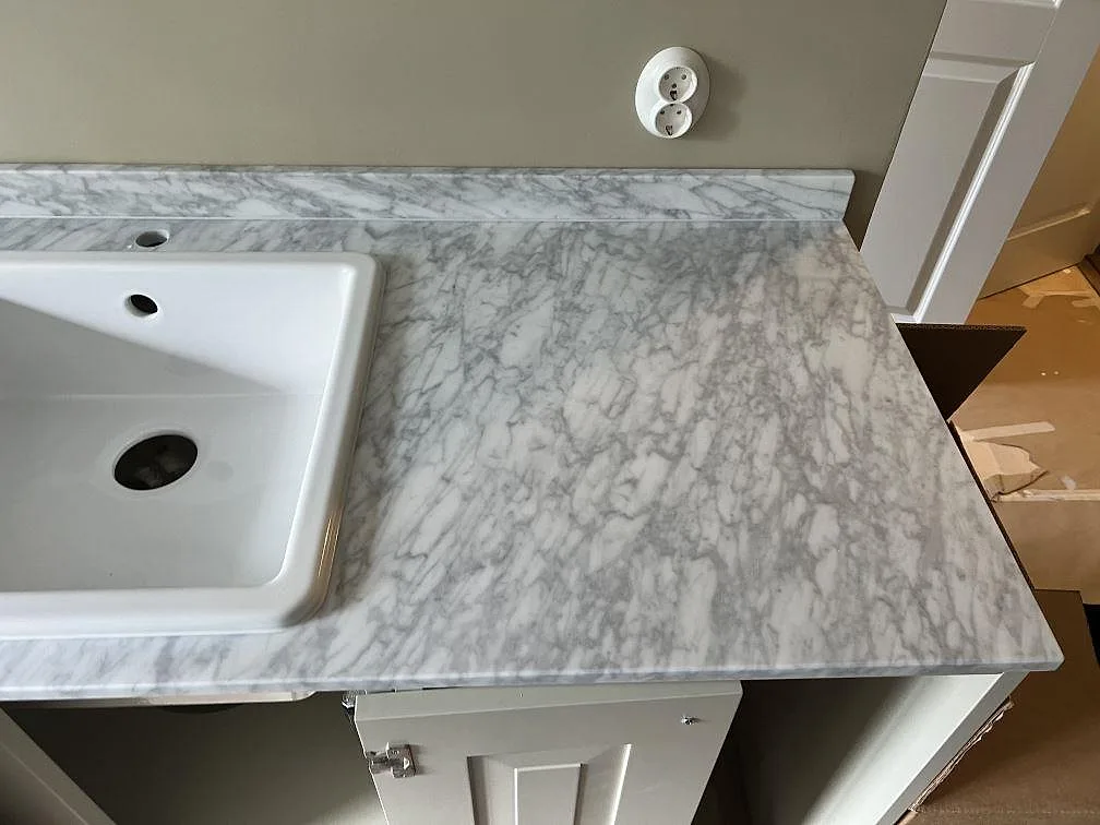 Detalj av matt Carrara C marmor i Malmö med vit fyrkantig diskho, blandarhål och låg stänkkant.