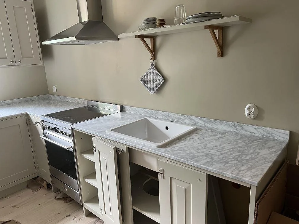 Köksbänkskiva i matt Carrara C marmor i Malmö med vit keramisk diskho, spiskåpa och krämfärgade luckor.